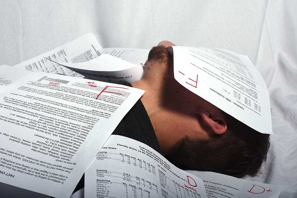 A student lays buried under a pile of white papers. The papers have big red letters on them ("F" and "D+") that show the student is failing their courses.