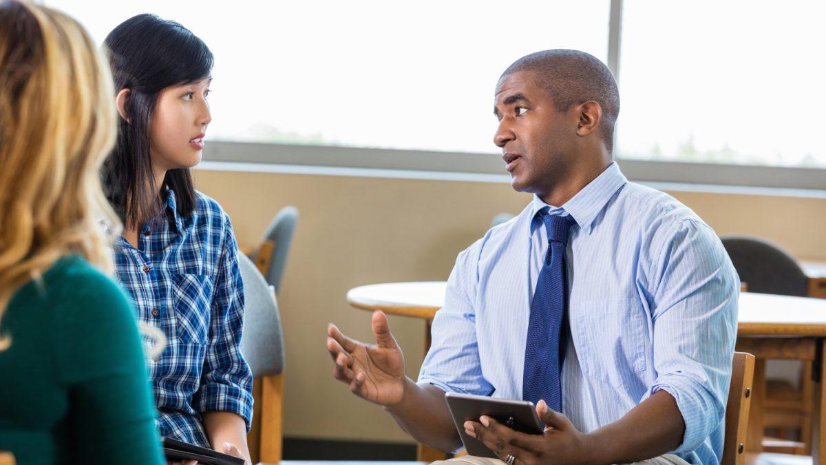 Mid adult African American male manager discuss current creative projects with employees in graphic design firm. He is gesturing as he talks about a project and is holding a digital tablet containing the details of the project. Diverse female employees are attentive as they listen to him. He is wearing a button down shirt with a blue tie.