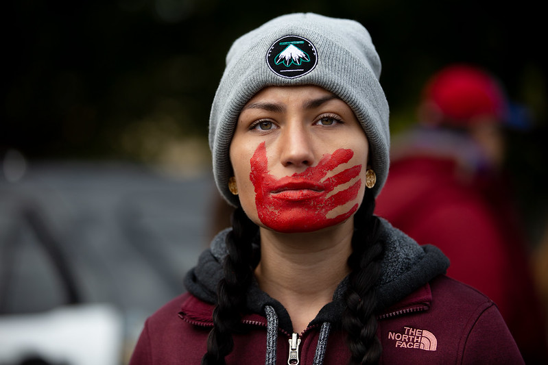 Image of a woman with a red hand print painted across her mouth, a symbol of the MMIW movement from Creative Commons.