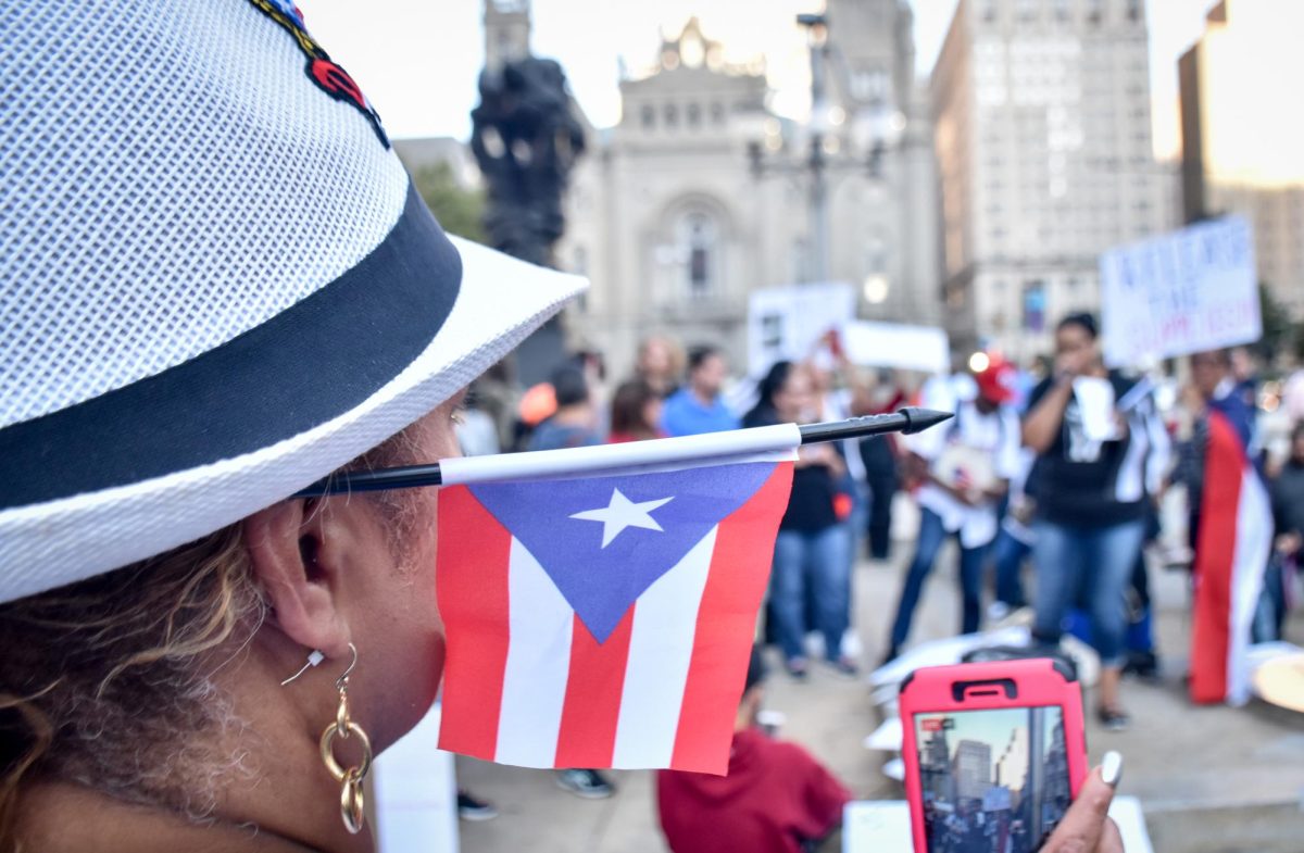 Image of a woman with a Puerto Rican flag behind their ear recording a political demonstration from Creative Commons